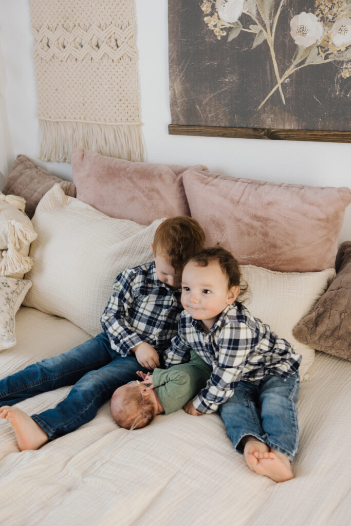 two toddler boys sit on a bed snuggling a newborn brother for professional family photos.