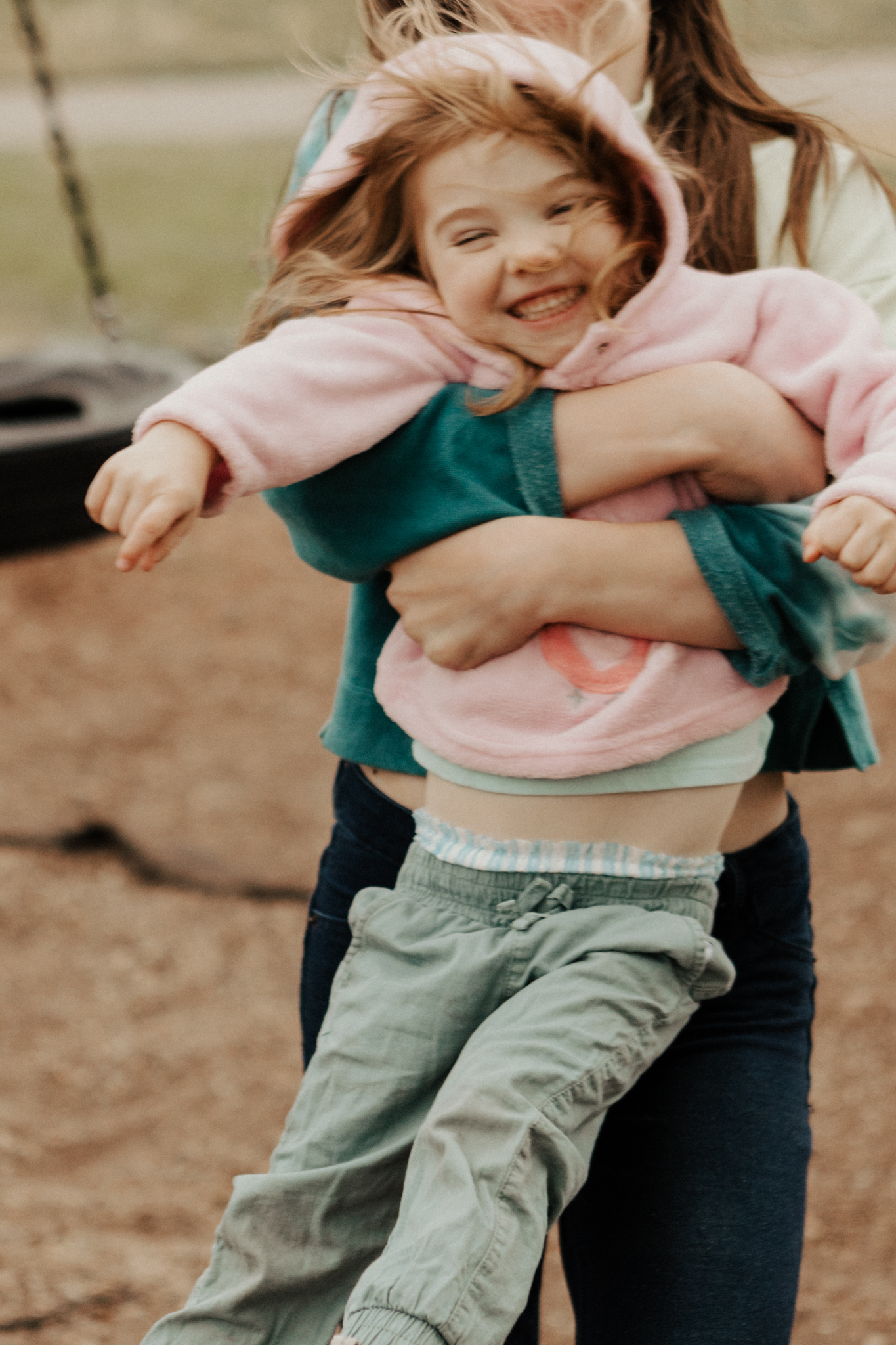A teenager and toddler sisters playing in the park. The older sister picks up and swings around the toddler and they both laugh with hair flying.