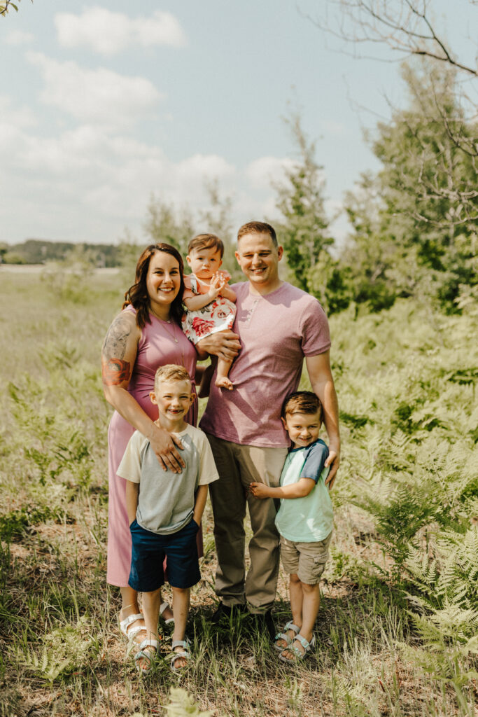 a father, mother, 2 school age boys and a 1 year old girl stand and smile together in a green field for family photos.